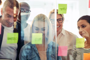 Diverse group of smiling business people standing in a modern office together brainstorming with sticky notes on a glass wall.