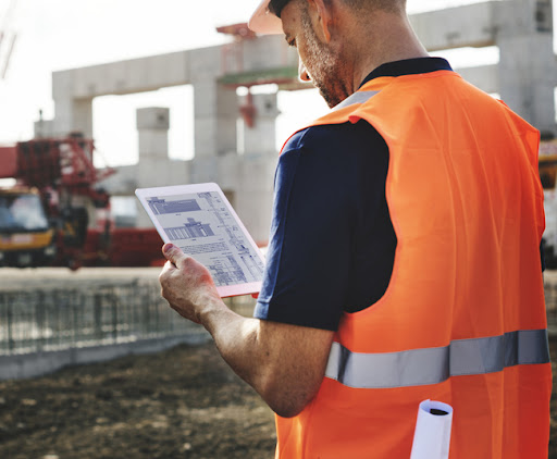 Male construction worker in a vest on a job site looking at plans on a tablet, and keeping up with technology trends.