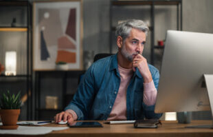 A business owner with gray hair wearing a denim button down works on his computer. Closely held business should review the IRS dirty dozen scams every year.