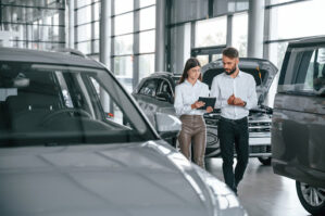 Two auto dealership employees walk between aisles of cars looking at a tablet. Dealerships can drive profitability through ancillary business.